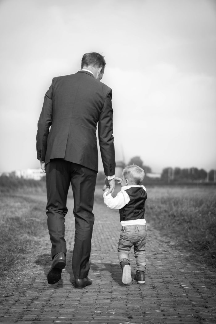 Older man in a business suit walks down a brick path while holding the hand of a young three-year-old boy.