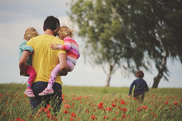 "Respectful parenting" father carries young children in his arms while another child runs into a field of flowers.